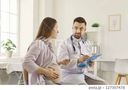 Male doctor sitting in medical office, listening to pregnant woman and discussing health concerns. Male doctor sitting in medical office, listening to pregnant woman and discussing health concerns. 127307478