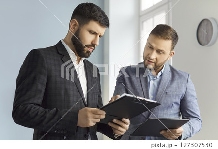 Portrait of two business men standing in office looking through financial documents. 127307530