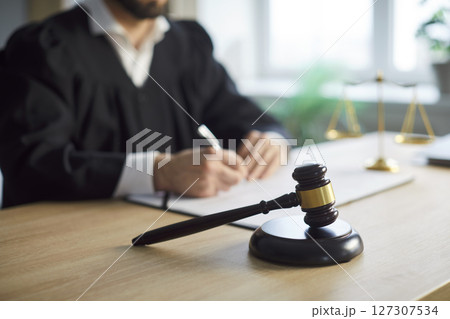 Judge passes fair judgement and rules, signing papers, posing at desk with gavel in foreground Judge passes fair judgement and rules, signing papers, posing at desk with gavel in foreground 127307534