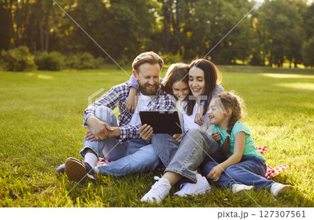 Happy family of four sitting on green grass in park and watching video on tablet during sunny day. 127307561