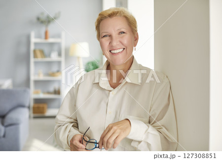 Portrait of mature woman standing in the living room and holding glasses in hand. Portrait of mature woman standing in the living room and holding glasses in hand. 127308851