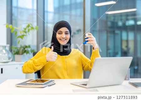 A smiling woman wearing a hijab in an office holds an inhaler, giving a thumbs up. Laptop and tablet are in the scene. 127311893