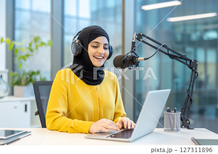 A smiling woman wearing a hijab hosts a podcast with a professional microphone and headset. She uses a laptop at a bright, modern workspace. A smiling woman wearing a hijab hosts a podcast with a professional microphone and headset. She uses a laptop at a bright, modern workspace. 127311896