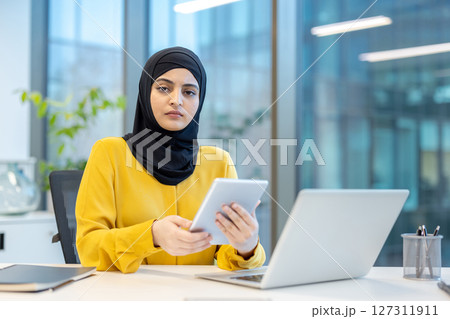 An Arab businesswoman in a yellow shirt and hijab works in an office, holding a tablet and using a laptop. An Arab businesswoman in a yellow shirt and hijab works in an office, holding a tablet and using a laptop. 127311911