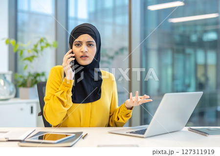 A woman wearing a hijab is on the phone, looking concerned while working on her laptop in the office setting. 127311919