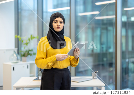 A woman wearing a black hijab and yellow top holds a tablet in a modern office setting with large windows. 127311924