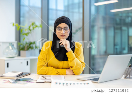 A Muslim businesswoman wearing a hijab sits at her desk in an office, looking at the camera with a serious expression. 127311952