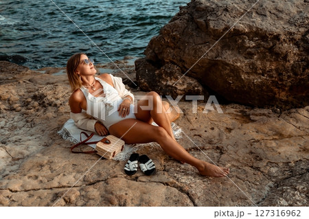 Woman Beach Relaxing - A woman in a white swimsuit and a white coverup relaxes on a beach with a rocky shore and ocean in the background. Woman Beach Relaxing - A woman in a white swimsuit and a white coverup relaxes on a beach with a rocky shore and ocean in the background. 127316962