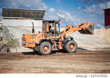 A loader is seen at a construction site, moving dirt and materials under a clear blue sky. Surrounding structures indicate ongoing building projects nearby. A loader is seen at a construction site, moving dirt and materials under a clear blue sky. Surrounding structures indicate ongoing building projects nearby. 127327050