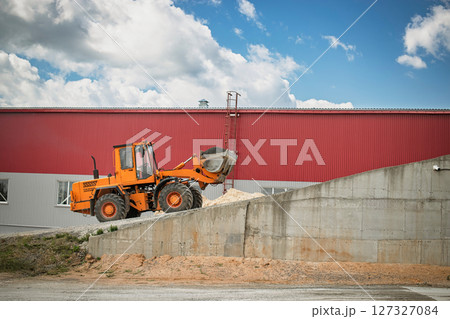 A construction vehicle moves gravel at a work site beside a large industrial building under a partly cloudy sky. A construction vehicle moves gravel at a work site beside a large industrial building under a partly cloudy sky. 127327084