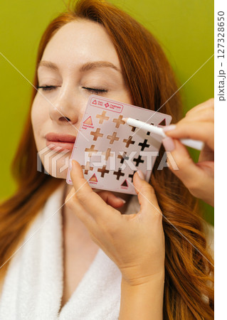 Vertical shot of cosmetologist holding markup grid on face of red-haired woman and marking with white pencil, preparing for beauty treatment on green background. Concept of beauty treatment. Vertical shot of cosmetologist holding markup grid on face of red-haired woman and marking with white pencil, preparing for beauty treatment on green background. Concept of beauty treatment. 127328650