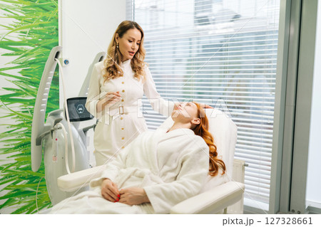 Skilled cosmetic professional doctor in white uniform discussing skin treatment options with female client lying on couch in modern beauty salon. Concept of non-surgery cosmetology, skincare. Skilled cosmetic professional doctor in white uniform discussing skin treatment options with female client lying on couch in modern beauty salon. Concept of non-surgery cosmetology, skincare. 127328661