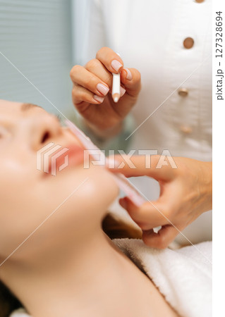 Vertical cropped shot of beautician preparing female client to facial cosmetic procedure, marking skin with white pencil and stencil, woman lying on treatment couch in beauty salon, close-up. 127328694