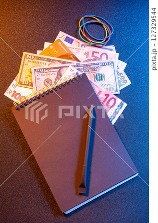 Assorted world currency with black notebook and pencil on dark background. 127329544