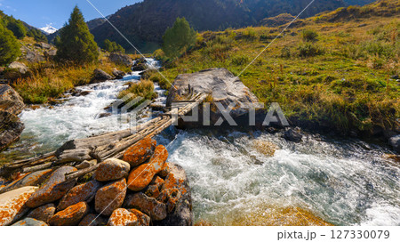Fast-flowing mountain stream with wooden bridge, rocky banks, and green vegetation under a blue sky. Fast-flowing mountain stream with wooden bridge, rocky banks, and green vegetation under a blue sky. 127330079