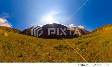 panoramic view of mountain valley with grassy slopes under a bright blue sky with scattered clouds. 127330082
