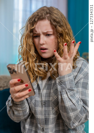 Young woman sits on sofa texting with anger waving hand annoyed by message using mobile phone chat 127334331