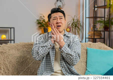 Young man touching cheek with expression of terrible suffer from painful toothache on sofa at home Young man touching cheek with expression of terrible suffer from painful toothache on sofa at home 127334401