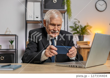 Elderly businessman playing game on smartphone exciting apps celebrate winning racing at home office Elderly businessman playing game on smartphone exciting apps celebrate winning racing at home office 127334404