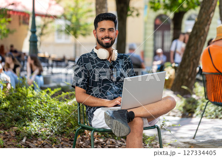 Indian man in headphones working online distant job with laptop browsing website chatting outdoors 127334405