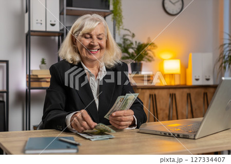 Excited senior businesswoman counting cash money dollar checking salary enjoy richness at workplace 127334407