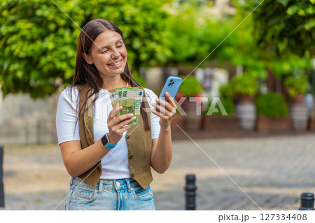 Happy Caucasian woman counting money euro cash, using smartphone calculator app on city street 127334408