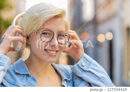 Portrait of smiling young woman in glasses taking off wireless headphones while looking at camera 127334420