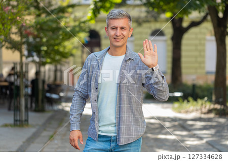 Cheerful mature man tourist smiling at camera, waving hands gesturing hello standing on street 127334628