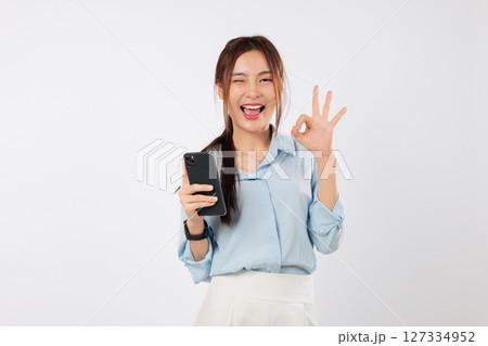 Excited Young Asian woman smiles, holding a mobile phone and giving an ok gesture sign. She is happy showing her smart phone. social media network isolated studio white background 127334952