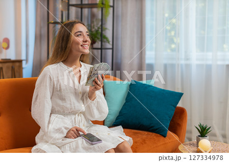 Rich happy young woman counting money cash using smartphone calculator sitting on sofa at home Rich happy young woman counting money cash using smartphone calculator sitting on sofa at home 127334978