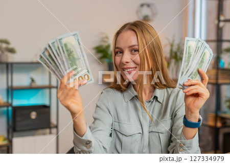 Excited businesswoman freelancer holding fan of dollar cash and dancing sitting at home office table Excited businesswoman freelancer holding fan of dollar cash and dancing sitting at home office table 127334979