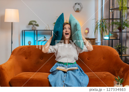 Irritated young woman reading book cover ears with pillows annoyed by loud noise sitting on sofa 127334983