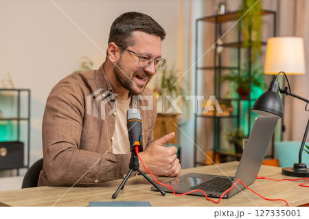 Happy young Caucasian man influencer speaking into microphone and laptop webcam at home office table Happy young Caucasian man influencer speaking into microphone and laptop webcam at home office table 127335001