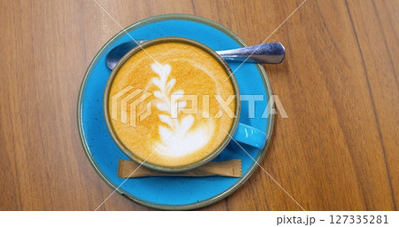 Creamy light brown latte art coffee with delicate foam pattern, wooden stirrer, metal spoon resting on blue ceramic saucer placed atop rustic wooden surface. Concept of coffee consumption 127335281