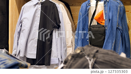 Fashion retail display presenting variety of clothing items, shirts, jeans, and black shoulder bag hanging neatly on store racks, showcasing contemporary casual menswear selection Fashion retail display presenting variety of clothing items, shirts, jeans, and black shoulder bag hanging neatly on store racks, showcasing contemporary casual menswear selection 127335303