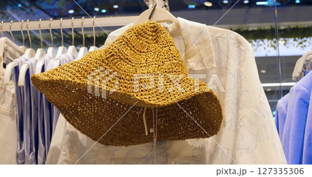 Woven straw sun hat hanging on clothing rack inside fashion retail store, showcasing summer accessory alongside shirts and other garments with natural, rustic texture. Concept fashion clothes 127335306