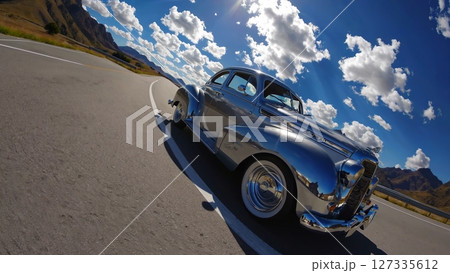 Polished chrome vintage car parked on a scenic mountain road gleams under the sunlight, reflecting the surrounding landscape and cloudy sky 127335612