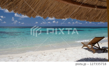 Pristine tropical beach featuring turquoise waters, white sandy shoreline, wooden sunbed positioned under straw umbrella with palm trees framing panoramic views of dreamy ocean and cloudy sky 127335718