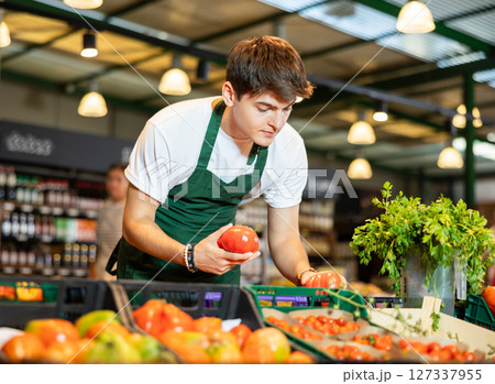 Skilled young seller arranging ripe red tomatoes on vegetable counter 127337955