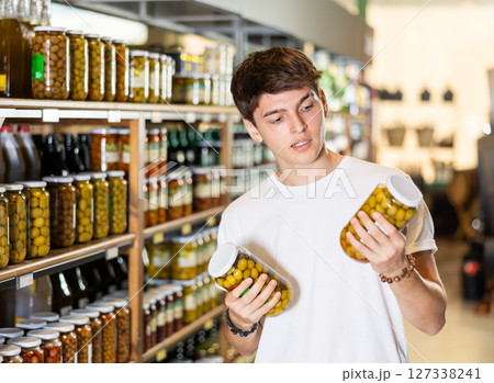 Young guy in supermarket selects examines compares two cans of olives. 127338241