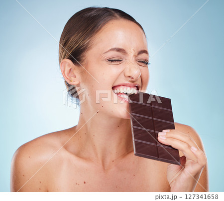 Skincare, beauty and woman with chocolate in a studio for a health, wellness and facial treatment. Cosmetics, diet and girl model eating sweet cacao candy while doing skin routine by blue background. Skincare, beauty and woman with chocolate in a studio for a health, wellness and facial treatment. Cosmetics, diet and girl model eating sweet cacao candy while doing skin routine by blue background. 127341658