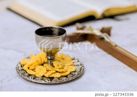 Silver chalice filled with beverage wine rests on plate of communion wafers near an open Bible near wooden cross. 127343336