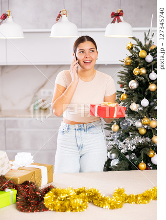 Cheerful young European woman in casual clothes enjoying mobile call near wrapped gifts and Christmas tree before holidays at flat 127345740