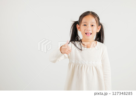Confident Asian young girl in a studio portrait isolated on white background makes a thumbs up OK sign, symbolizing agreement and success. kindergarten child smile radiates positivity and satisfaction 127348285