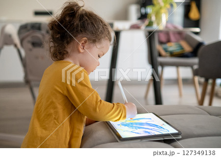 Small girl focused on tablet screen, drawing with pencil in a bright and tidy living room. Technology and creativity at home. Small girl focused on tablet screen, drawing with pencil in a bright and tidy living room. Technology and creativity at home. 127349138