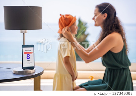 Mother puts sun hat on daughter as smartphone displays high UV alert. Practical response to sun safety warning for child protection 127349395