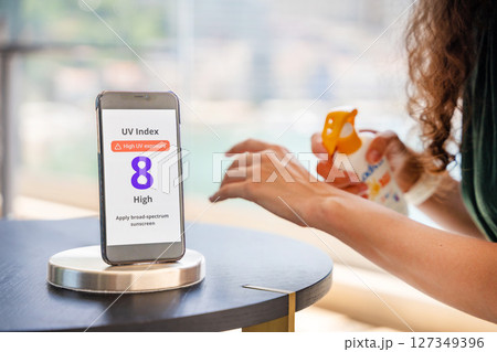 Smartphone with high UV alert in focus, while woman in background applies sunscreen to her skin. Everyday sun protection routine in response to radiation warning. Smartphone with high UV alert in focus, while woman in background applies sunscreen to her skin. Everyday sun protection routine in response to radiation warning. 127349396