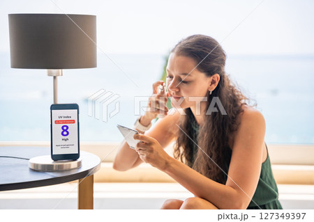 Smartphone with high UV alert in focus, while woman applies sunscreen to her face in mirror. Conscious skincare routine inspired by sun safety warning. 127349397