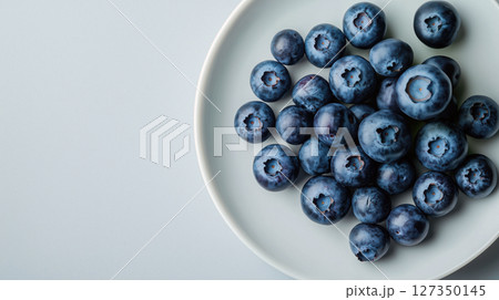 Fresh ripe blueberries on a white plate with soft blue background. Minimalist composition emphasizes natural texture and color. Top view. 127350145