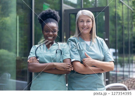 Smiling female doctors in lab coats standing near clinic and looking confident 127354395